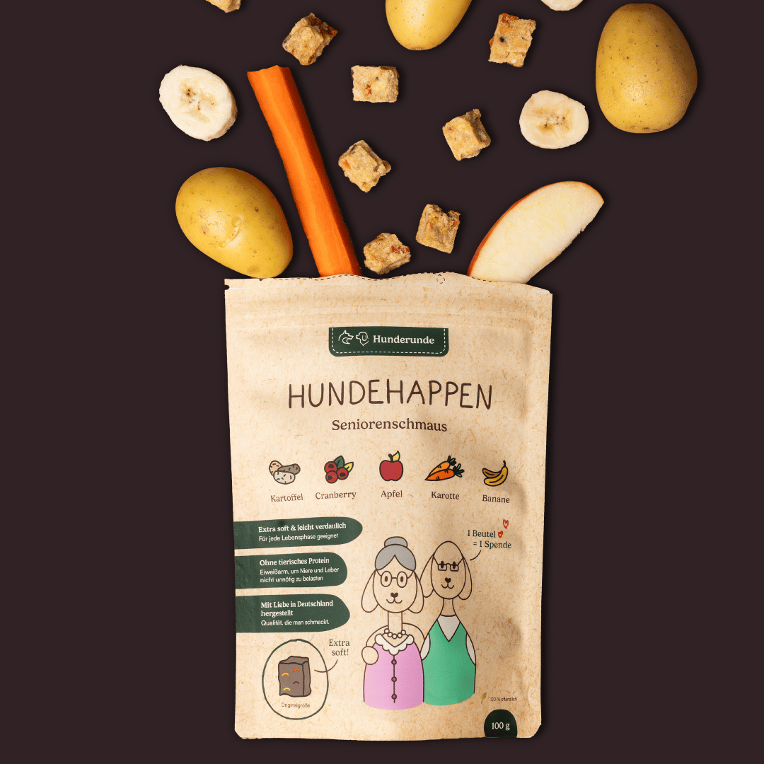 Hunderunde Hundehappen Seniorenschmaus Leckerli-Tüte mit Zutaten (Kartoffel, Karotte, Banane, Apfel) und Leckerli-Würfeln auf dunklem Hintergrund.