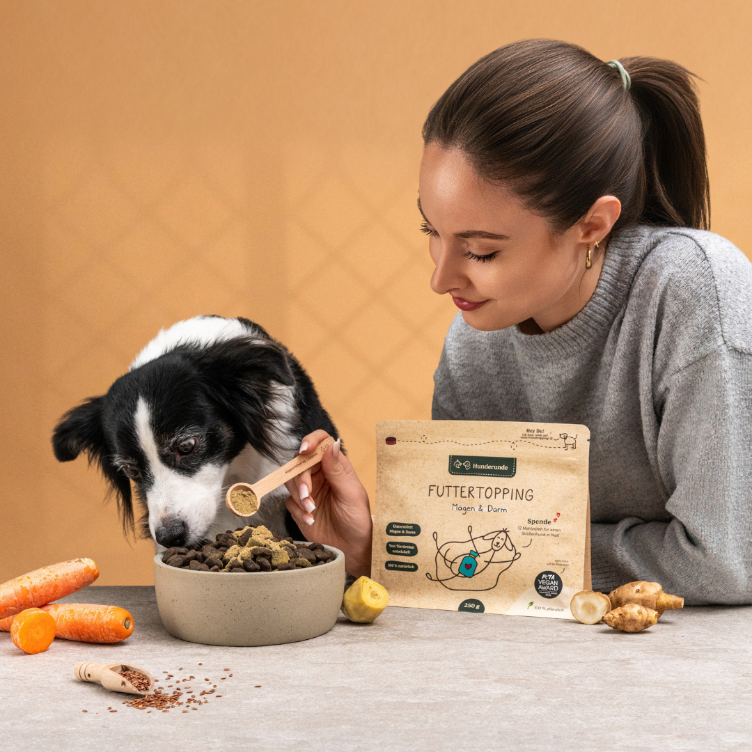 A woman uses the Portionierer, which holds about 1g of topping, to add powder to her dog's food while the dog eats; veggies and a food package are on the table.