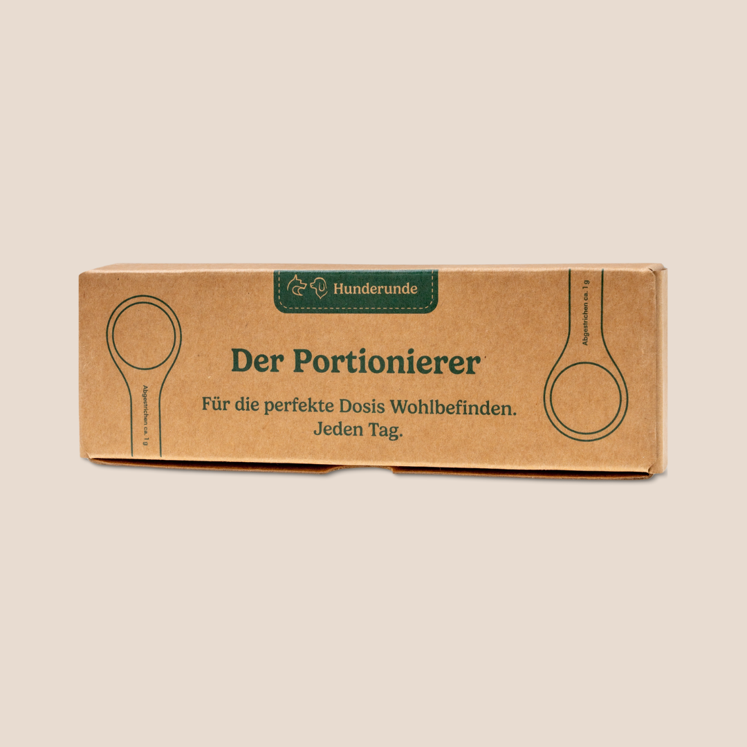 Brown cardboard box labeled "Portionierer - fasst ca. 1 g vom Topping" with green text and spoon illustrations on a beige background.