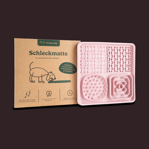 Die Schleckmatte ist eine rosa Leckmatte für Hunde mit unterschiedlichen Oberflächenstrukturen, die neben ihrer Kartonverpackung mit der Aufschrift „Schleckmatte“ abgebildet ist.